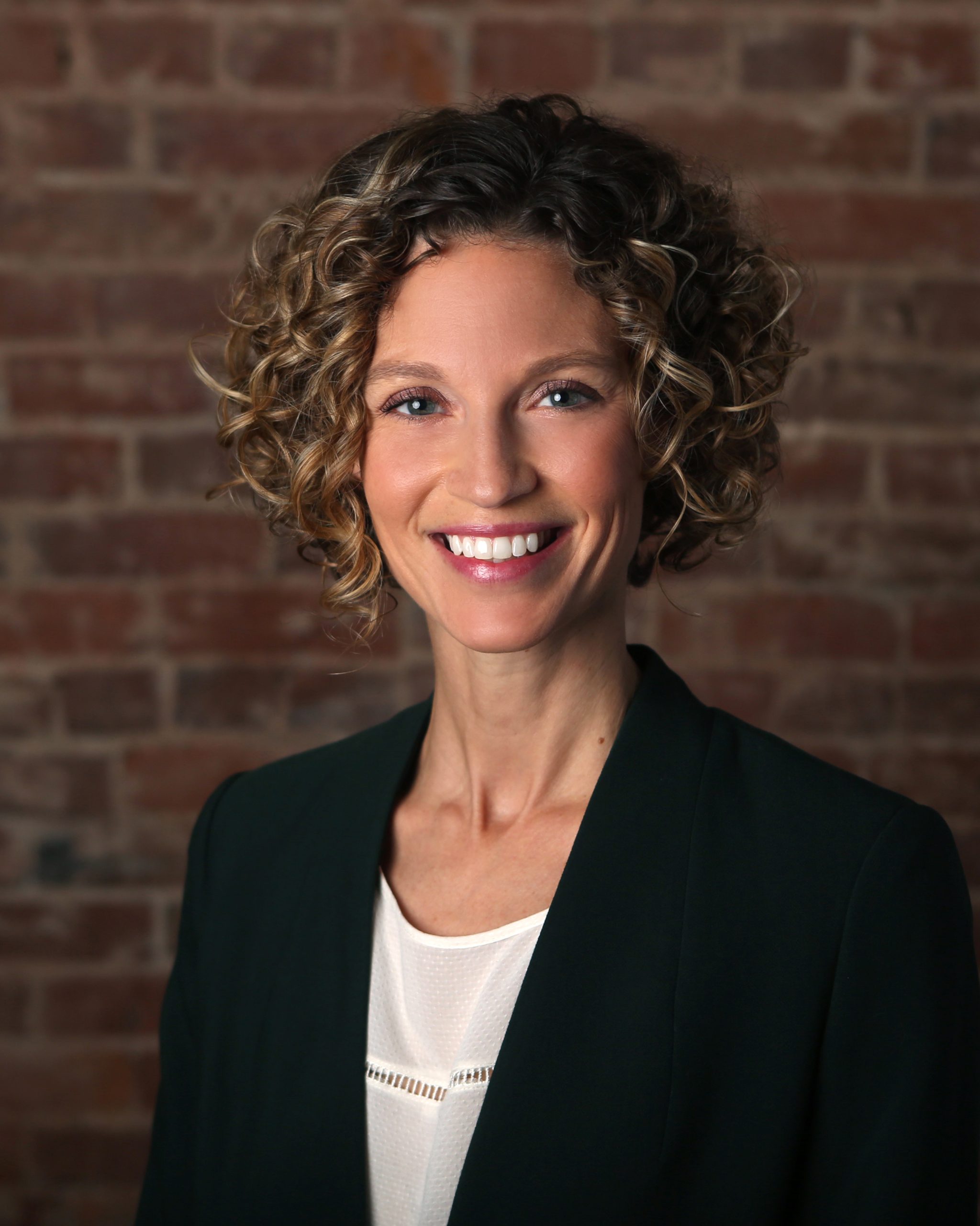 Headshot of smiling woman in dark jacket and white shirt against a brick background