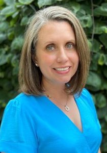 Photo of Camille Gill - she is smiling at the camera and wearing a blue shirt, standing in front of a plant.