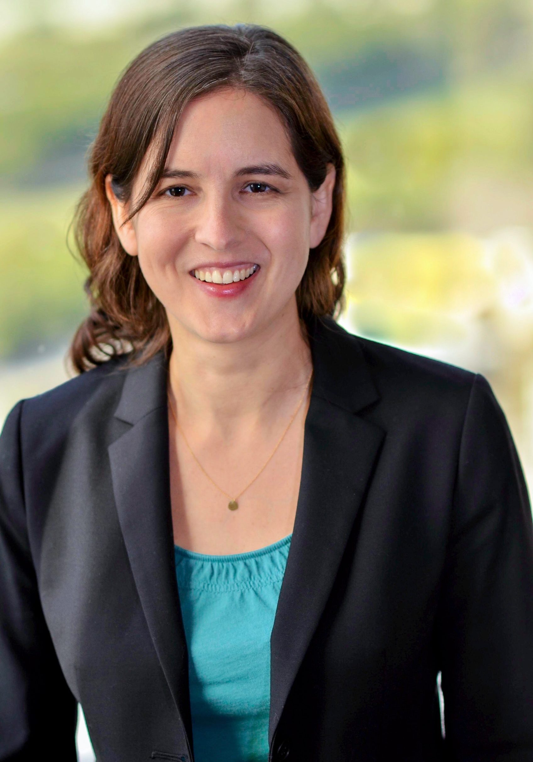 Headshot of smiling woman in black jacket and teal shirt in front of outdoor background