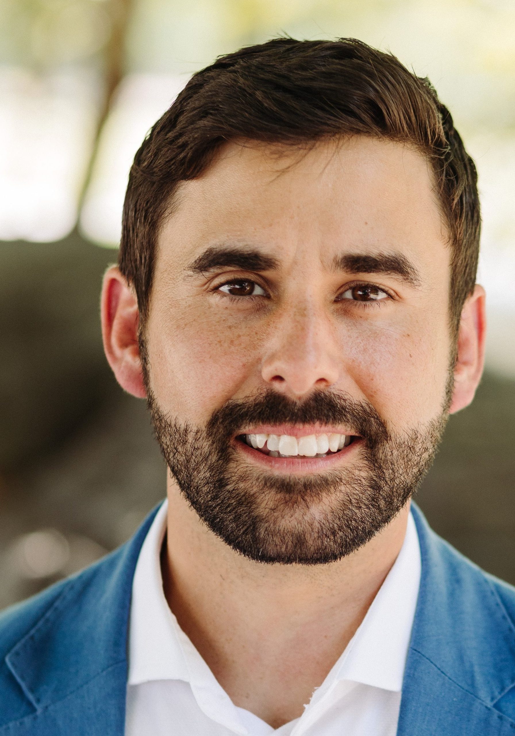 Headshot of man smiling in blue jacket and white shirt in front of outdoor background