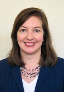 Headshot of smiling woman in blue jacket with white shirt and blue necklace against a tan background